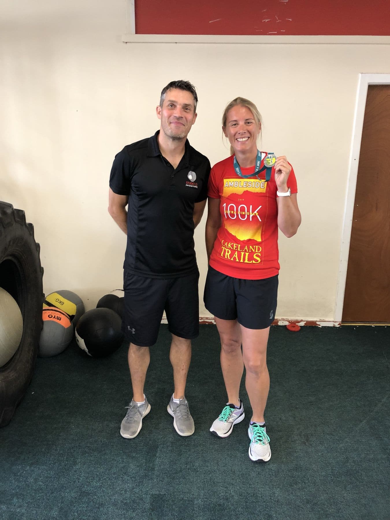 Runner in her 100K ultra finisher tee showing her medal with Richard Adams at Studio 121